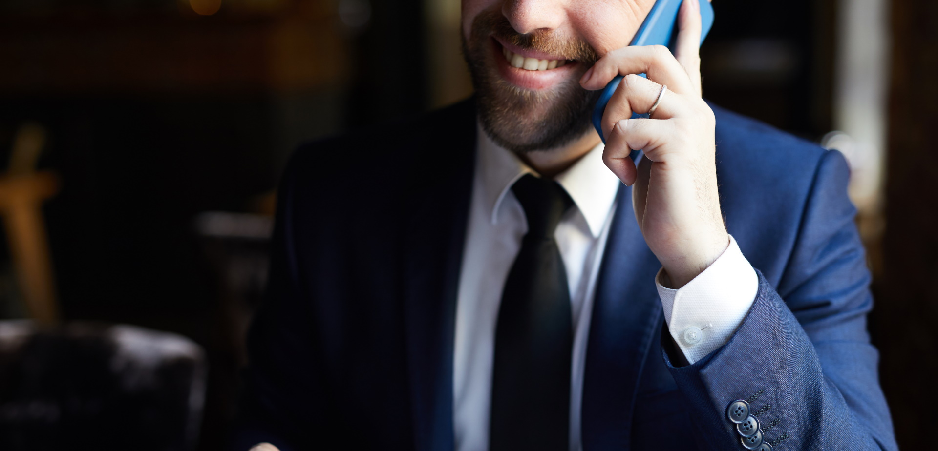 positive-businessman-with-coffee-calling-on-phone-2MZNE9B Close-up of positive bearded businessman in elegant suit with coffee cup calling on mobile phone and smiling while sitting at table